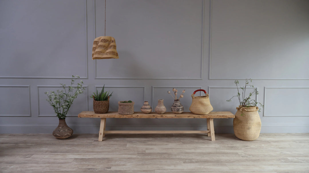 A collection of hand woven baskets and lamp shades on a rustic wood bench with flowers and foliage