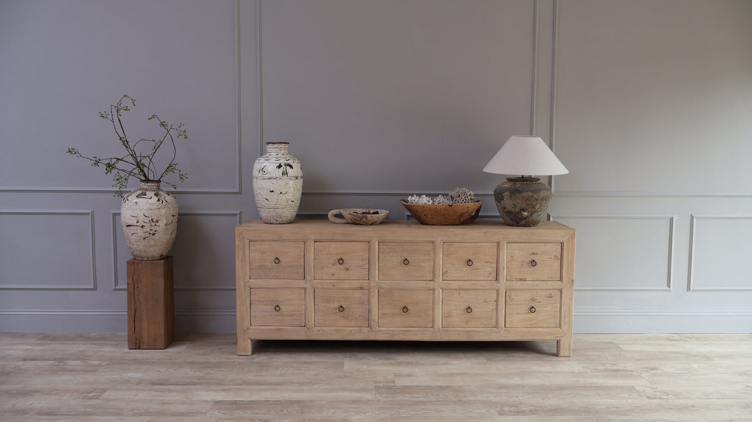 A large rustic stone table lamp displayed on a Home Barn pale reclaimed wood cabinet together with a selection of antique urns and bowls