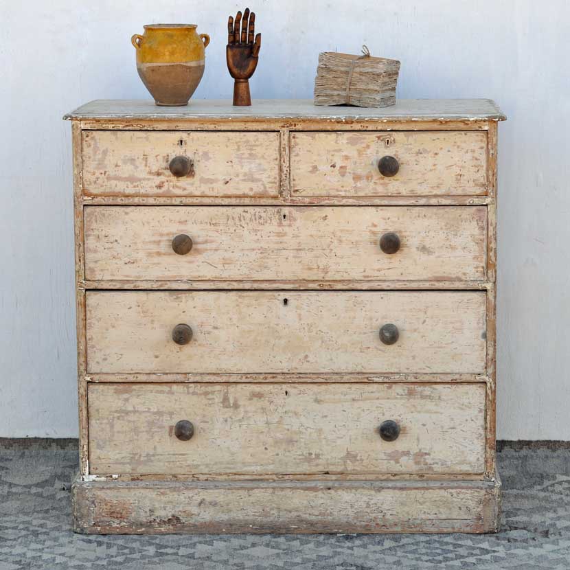 A painted and distressed vintage chest of drawers displaying an antique French confit pot, vintage book bundle and a wooden artists hand against a plain white backdrop