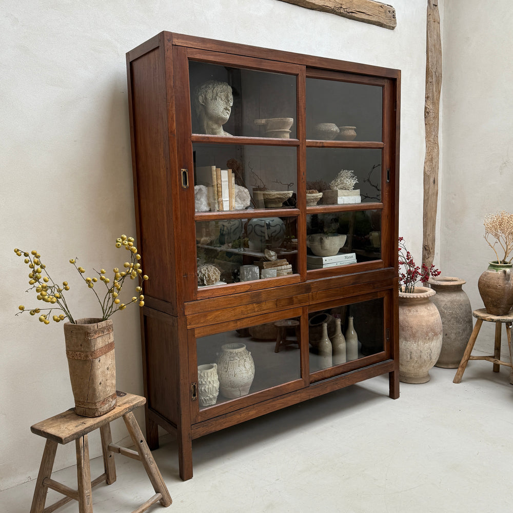Wooden display cabinet with glass doors filled with decorative items, surrounded by vases and plants.