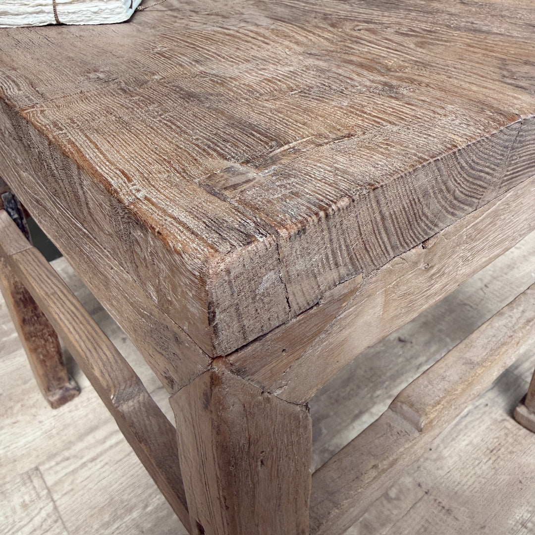 Close-up of a rustic wooden table corner showing the thick reinforced wood joint and detailed grain texture, with a light wood floor visible underneath.