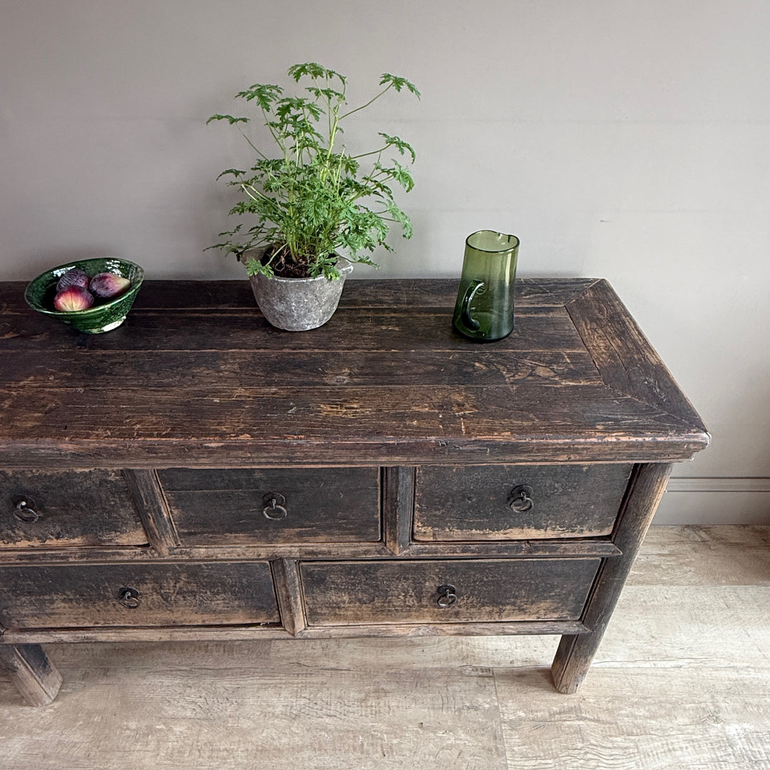 A dark rustic wooden sideboard or dresser with distressed brown and black weathered paint, styled with a potted green plant, a bowl of apples, and a green glass vase against a plain light gray wall.
