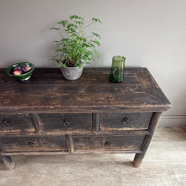 A dark rustic wooden sideboard or dresser with distressed brown and black weathered paint, styled with a potted green plant, a bowl of apples, and a green glass vase against a plain light gray wall.