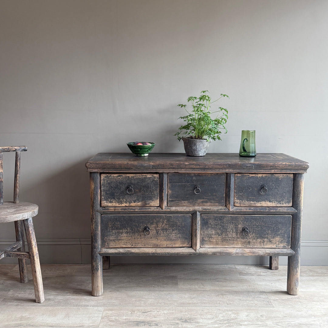 A rustic dark wood chest of drawers with six drawers, styled with a potted plant, a green bowl, and a green glass vase against a neutral gray wall.