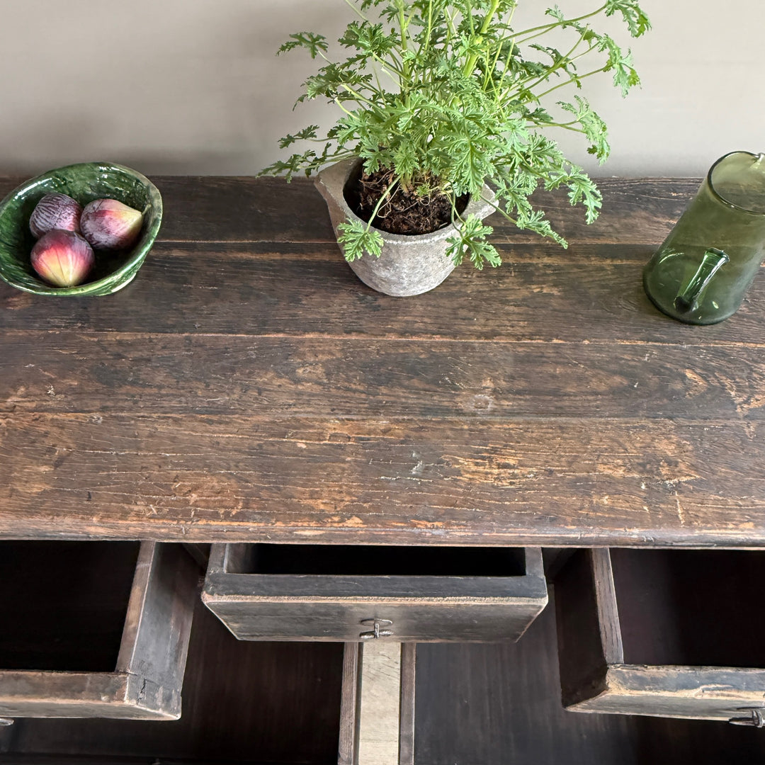 A rustic dark brown wooden console table with distressed finish, featuring an open drawer, styled with a potted green plant, a bowl of figs, and a green glass pitcher against a neutral wall.