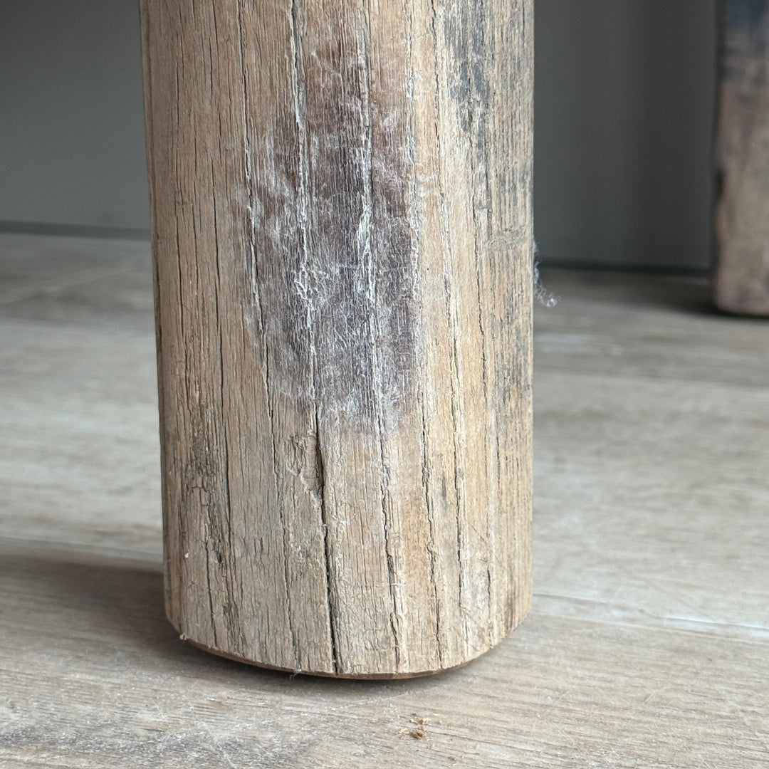 A close-up of a cylindrical wooden post or leg with a weathered, rustic finish and visible grain, standing on a light-colored floor.