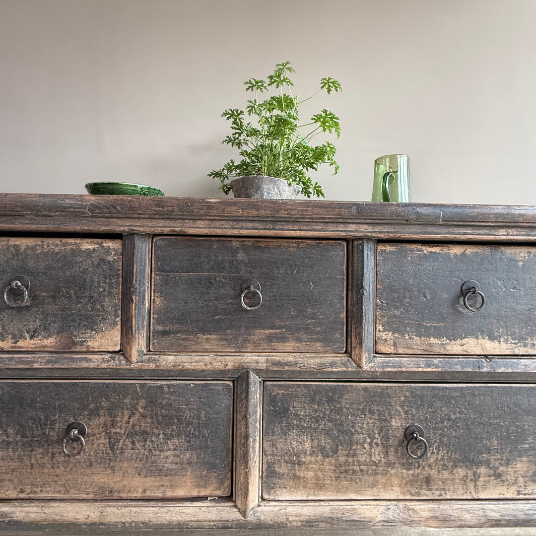 A rustic dark brown wooden dresser with six drawers and distressed metal ring pulls, styled with a potted green plant and a green glass mug on top against a plain beige wall.