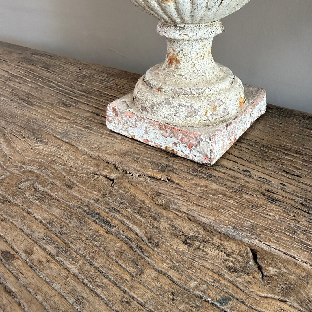 A close-up of a distressed, vintage-style white stucco urn base with peeling paint, resting on a rustic, weathered wooden surface.