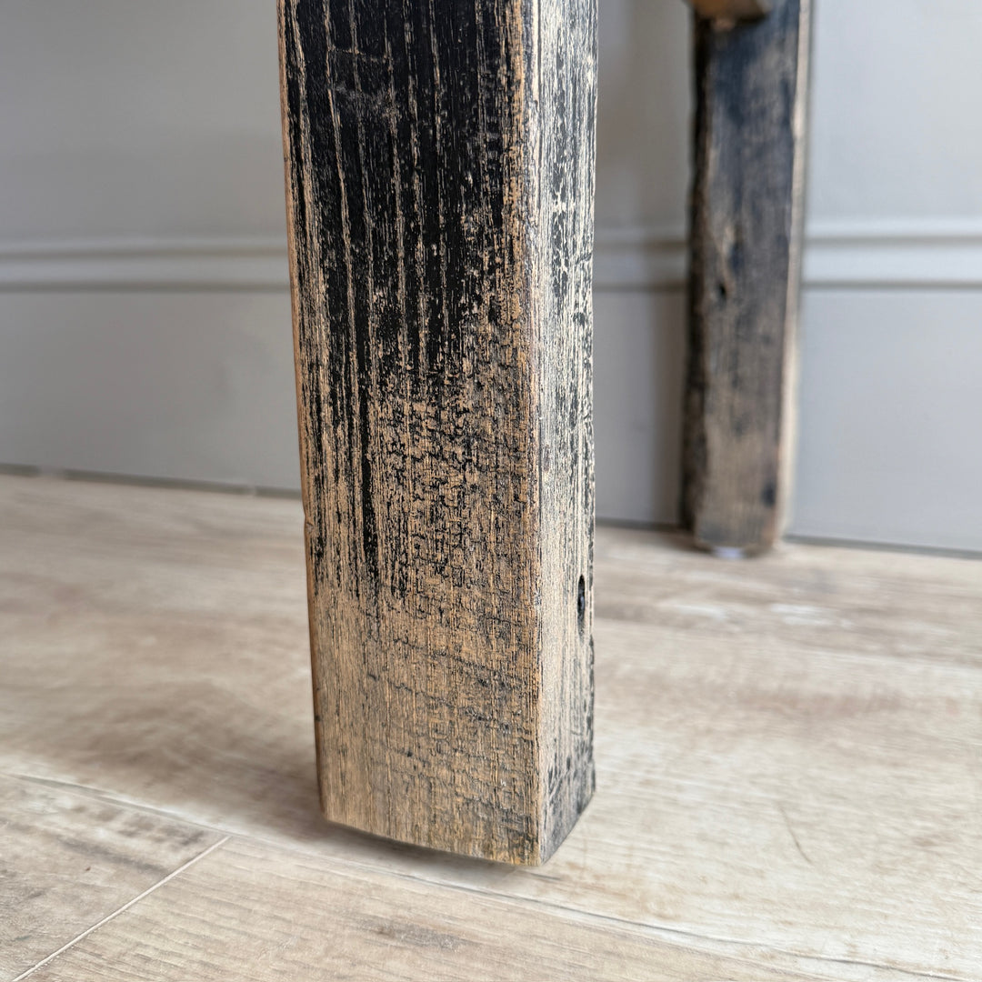 A close-up of a rustic dark wood table leg with a distressed black and brown finish, standing on a light wood floor against a gray wall.