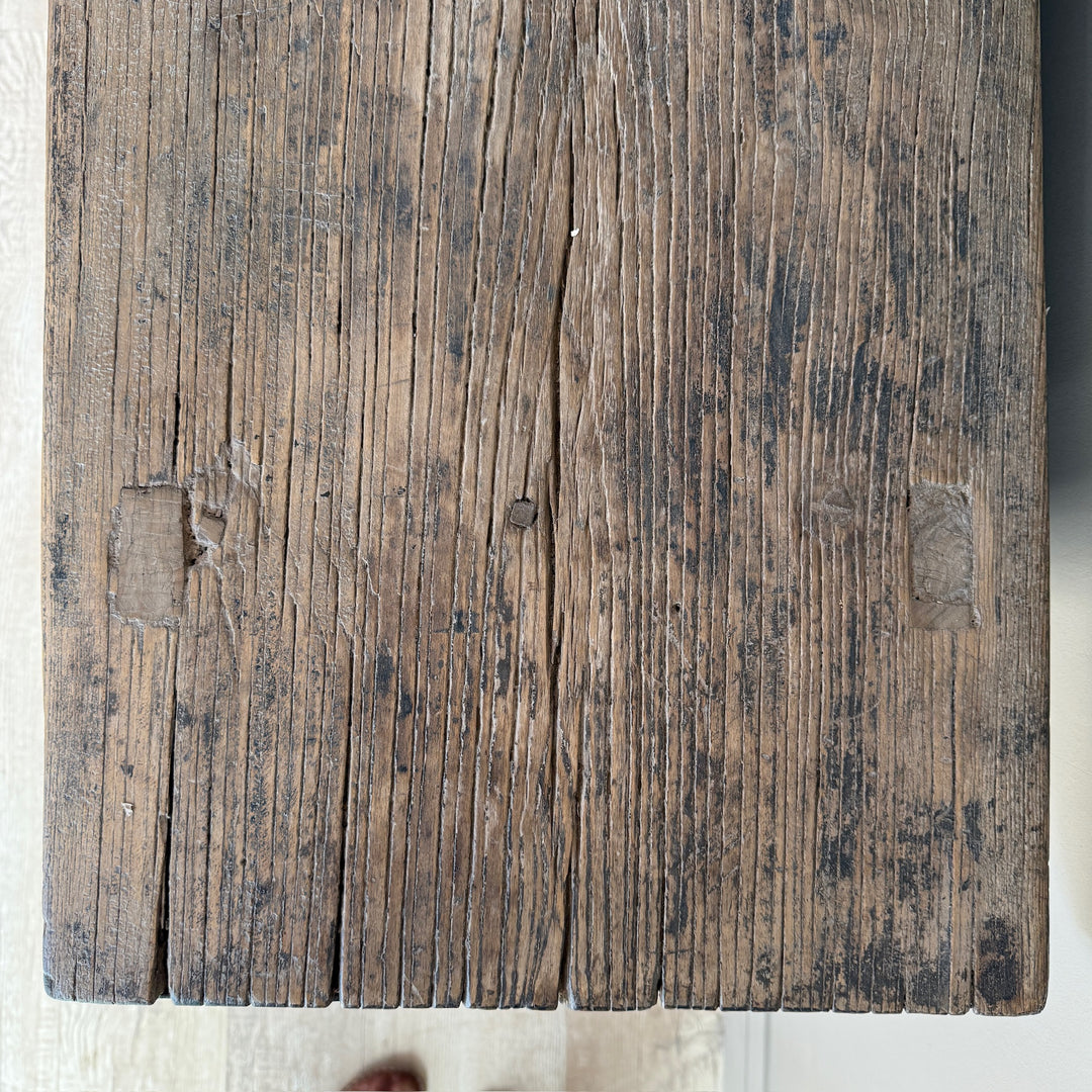 A close-up of a rustic, weathered wooden plank surface with prominent grain, cracks, and darkened areas, showing a light-colored floor underneath.