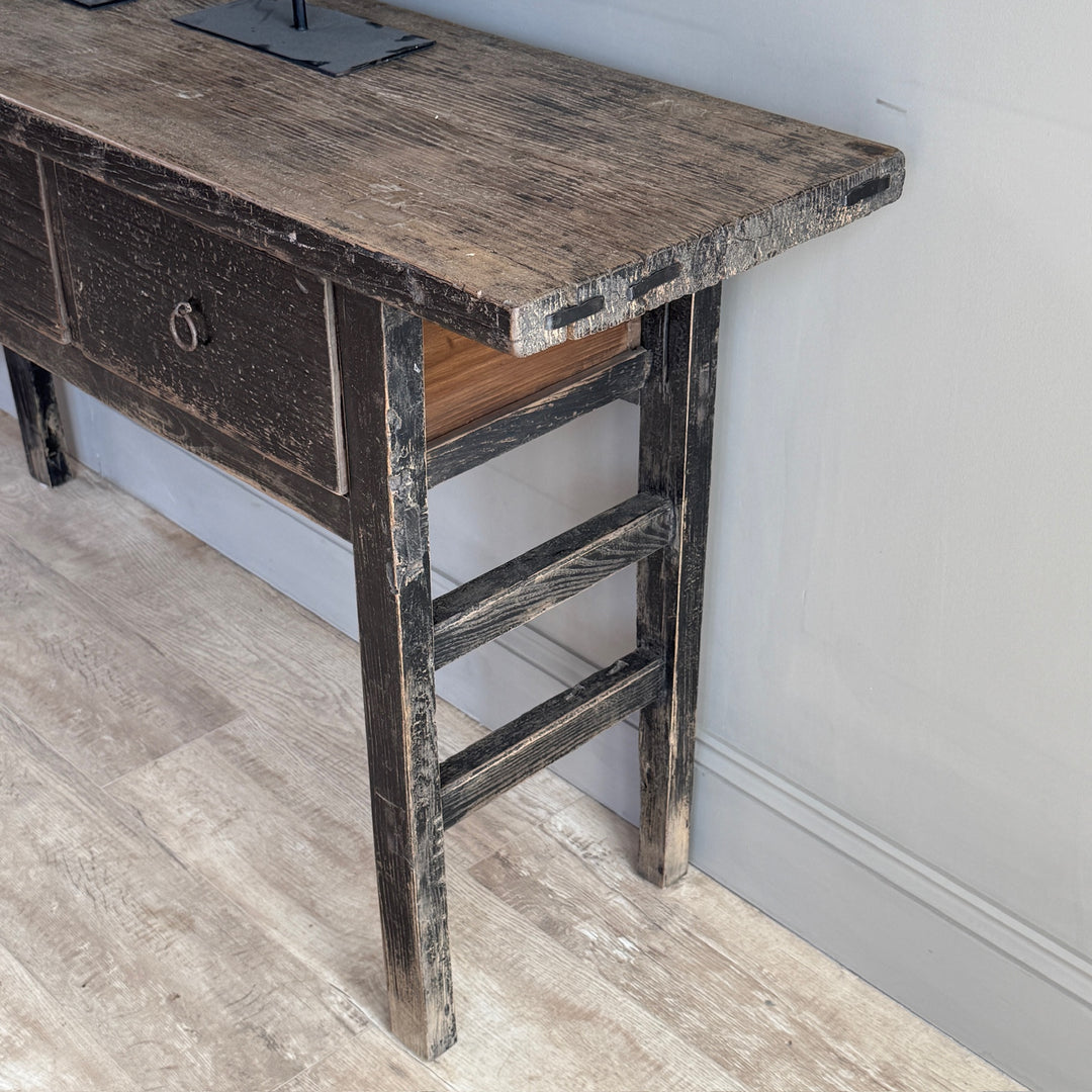A rustic dark wood console table with a distressed finish, featuring a single drawer with a metal pull handle and open lower storage compartments, shown in a home interior setting.