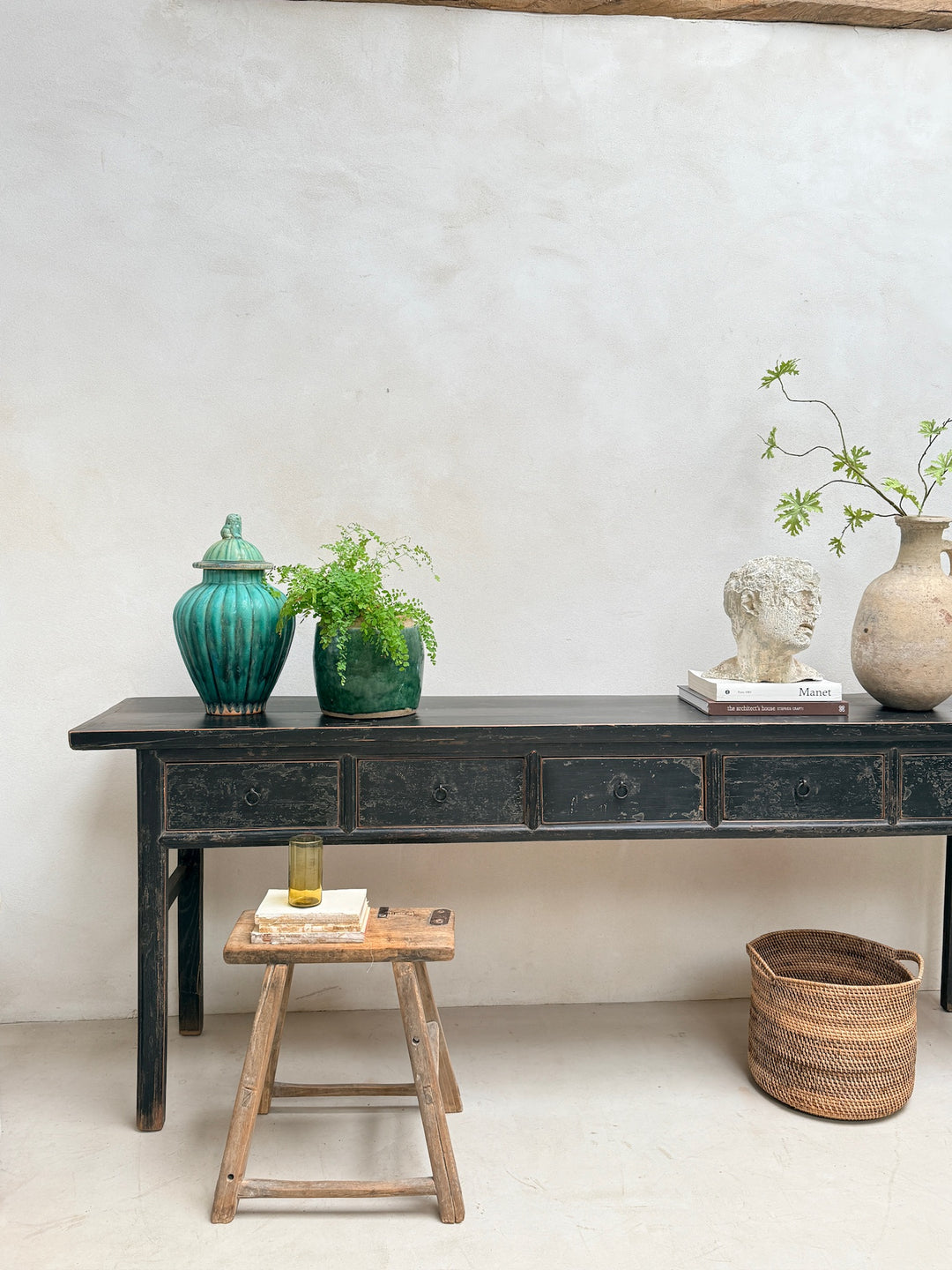 A long black wooden console table with drawers, styled with a teal ceramic jar, a green potted plant, a white bust sculpture, a beige vase with leafy stems, a small wooden stool, and a woven basket against a textured off-white wall.