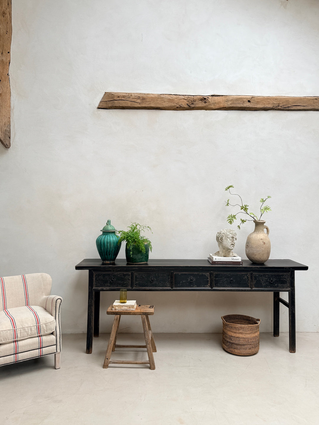 A dark wooden console table styled in a minimalist room with a textured plaster wall, a rustic wooden beam, a striped armchair, and decorative ceramic vases and plants.
