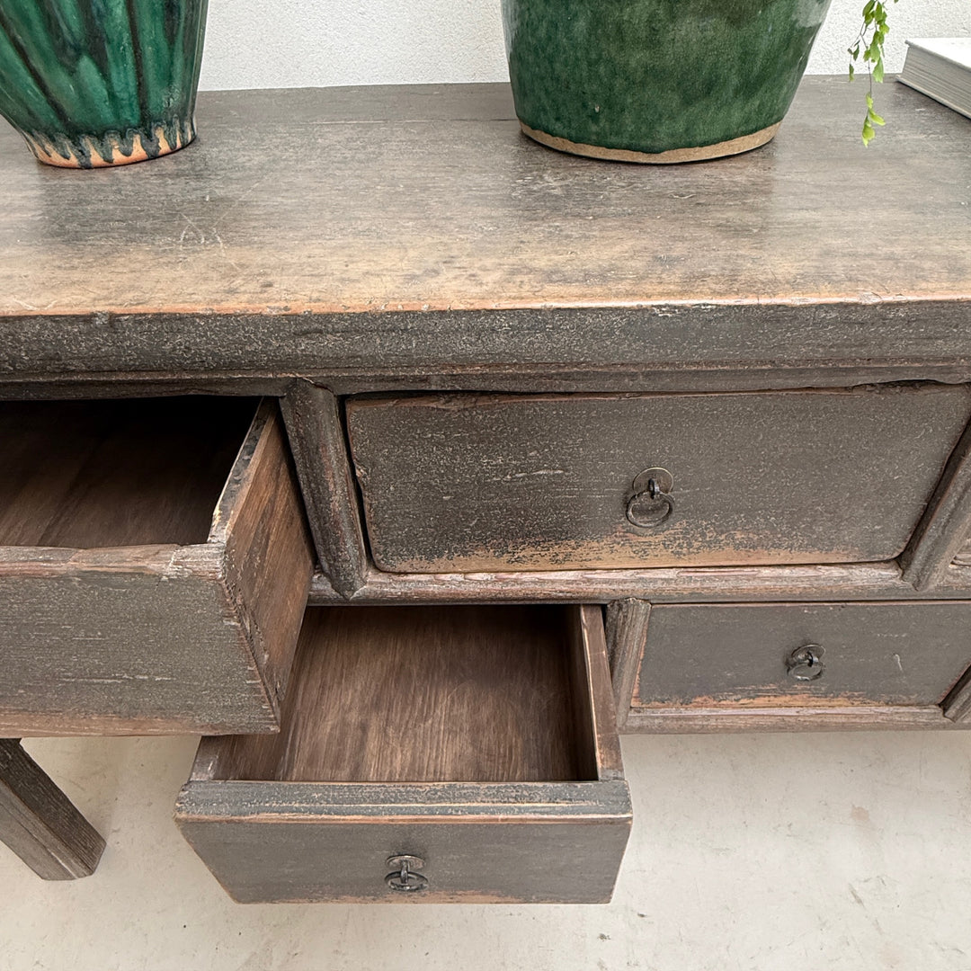 A close-up of a distressed dark brown wooden writing desk with multiple drawers, showing one open drawer. Two green ceramic plant pots sit on the wooden tabletop against a plain white wall.