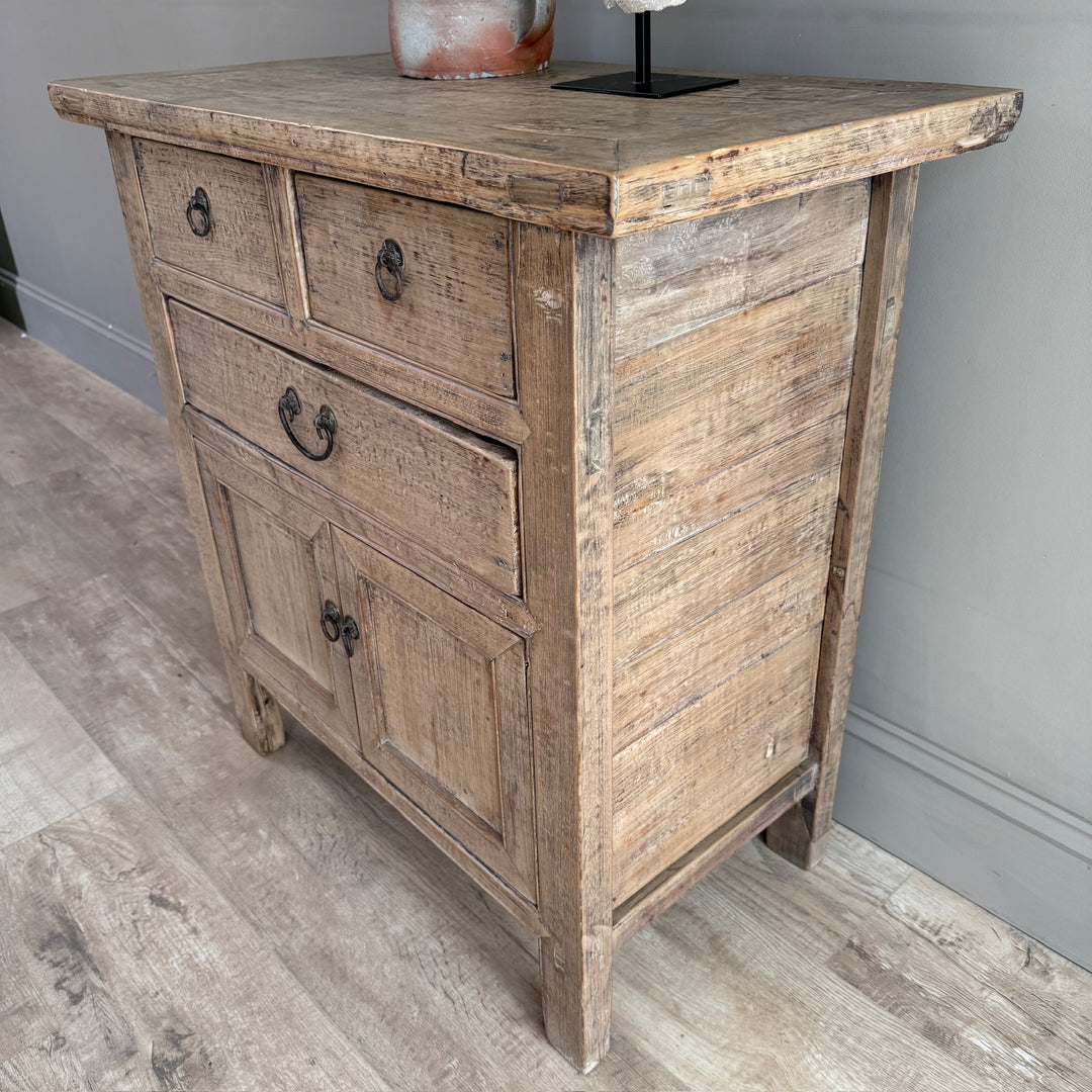 A rustic, weathered wood sideboard cabinet with three drawers on top and two doors below, featuring dark metal handles, shown on a light wood floor against a gray wall.