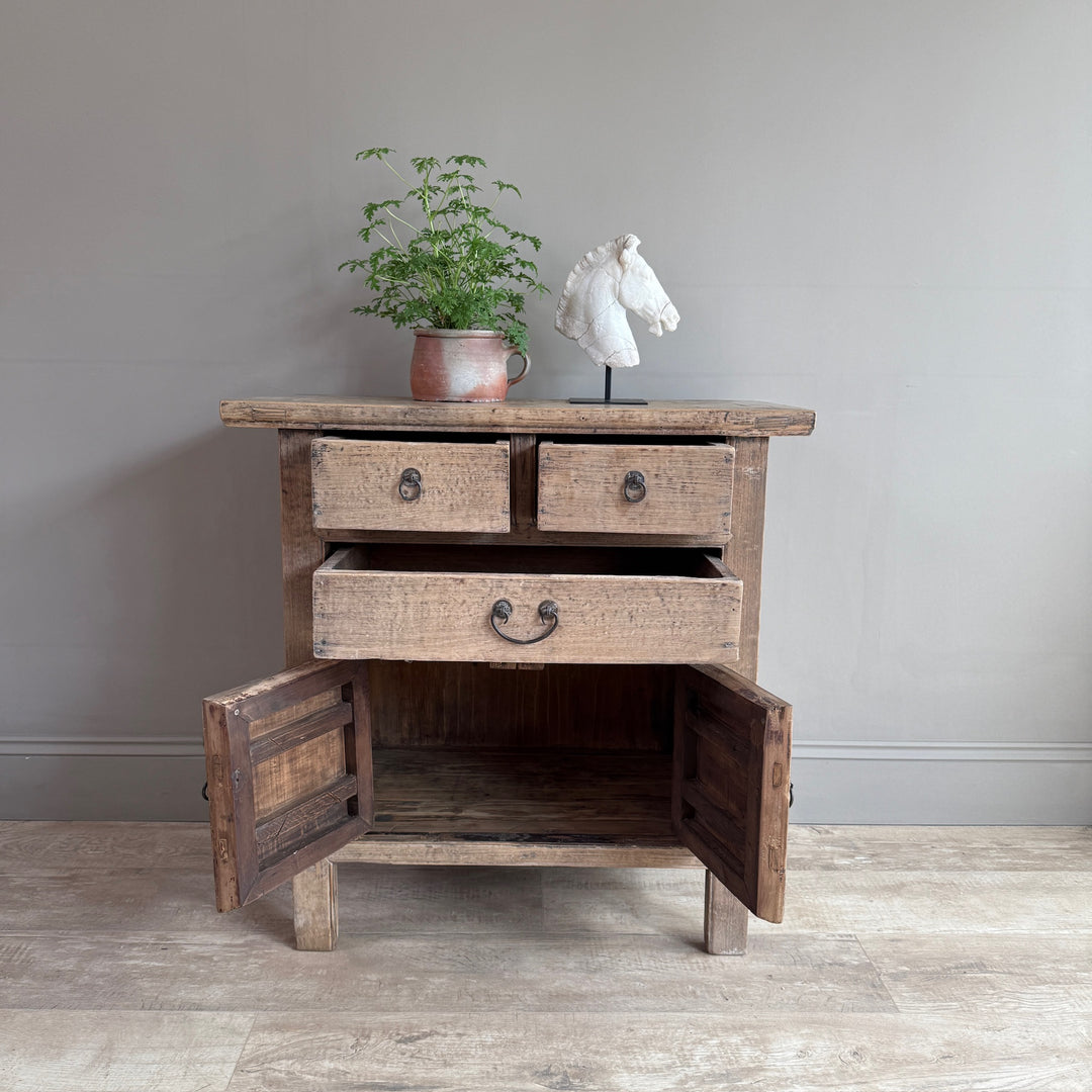 A rustic wooden side cabinet with two drawers on top, one open drawer in the middle, and two open doors below, shown against a plain gray wall on a light wood floor. A potted green plant and a white horse bust sculpture sit on top of the cabinet.