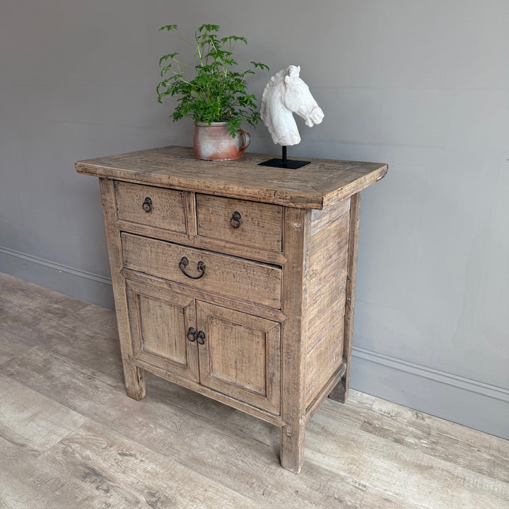 A rustic light brown wooden sideboard cabinet with three drawers and two lower doors, styled with a potted green plant and a white horse head sculpture on a gray wall and wood floor.