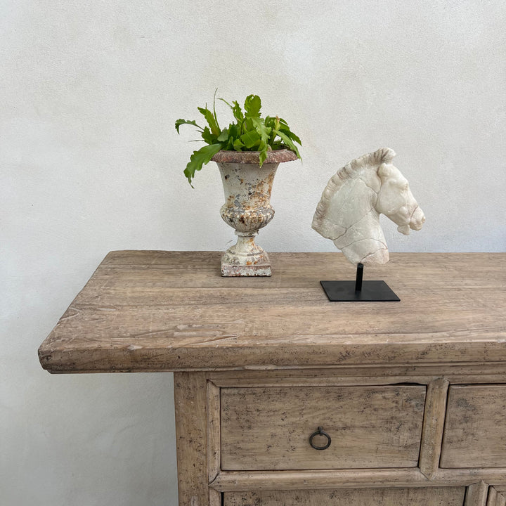 Wooden console table with decorative items including a plant in an urn and a horse head sculpture.