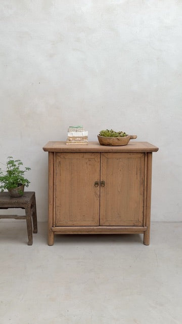 A rustic light brown wooden cabinet with two doors and small round metal knobs, styled with a small stack of books and a bowl of greenery on top, set against a textured light gray wall.