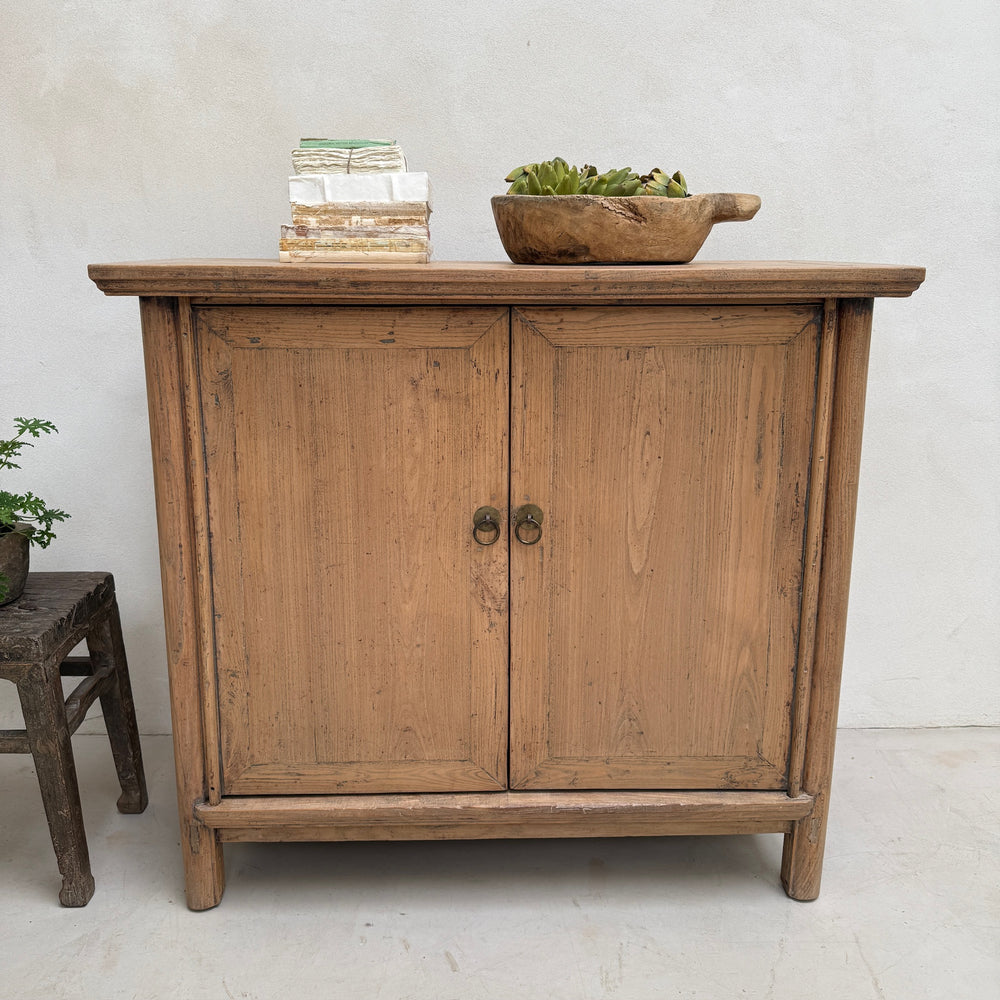 A rustic wooden cabinet with two doors and round metal knobs, styled with books and a bowl of greens on top, set against a light textured wall.