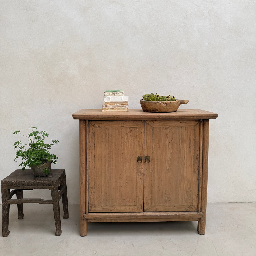 A rustic light brown wooden cabinet with two doors and small round metal knobs, styled in a minimalist room with a textured off-white wall. A small wooden stool with a potted green plant sits to the left, and a wooden bowl with greenery is placed on top of the cabinet.