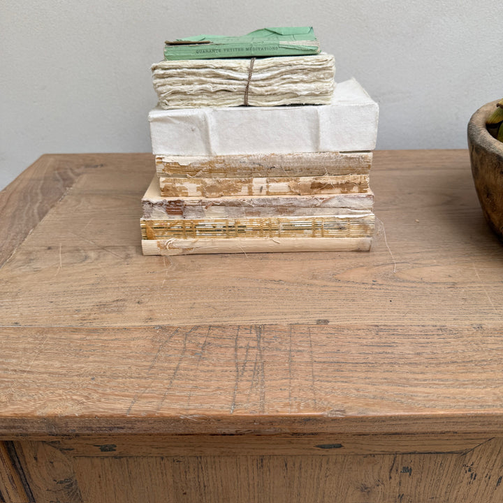 A stack of vintage books and stacked paper bundles on a rustic wooden table against a plain light gray wall.