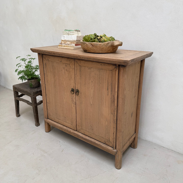 A rustic light brown wooden sideboard cabinet with two doors and dark metal knobs, styled with a bowl of greenery and books on top, next to a small dark wooden stool against a white wall.