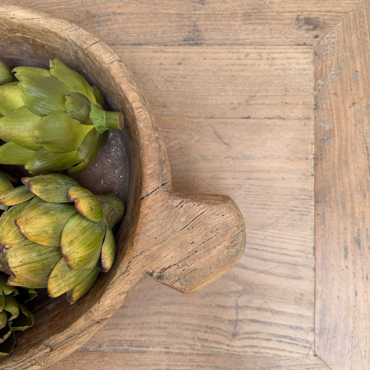 Green artichokes in a rustic wooden bowl on a light wood tabletop.