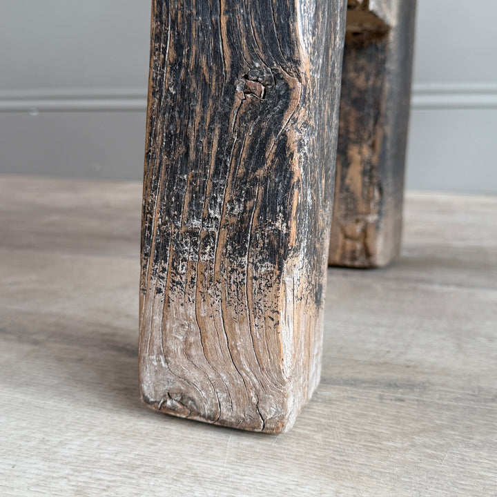 A close-up of a rustic wooden furniture leg with a heavily weathered, dark-charred top and light-toned lower section, standing on a light wood floor.