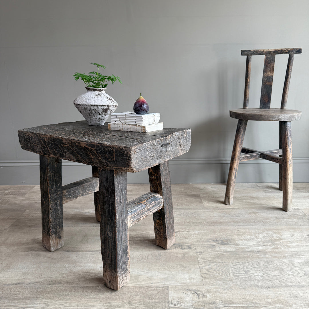 A rustic dark wood stool with a thick rectangular seat and crossed front legs stands in a room with light wood flooring and a gray wall. A small potted plant and stacked books with a fig on top sit on the stool, while a second matching wooden chair is visible in the background.