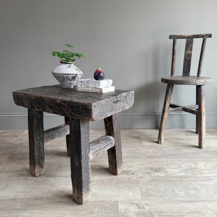 A rustic dark wood stool with a thick rectangular seat and crossed front legs stands in a room with light wood flooring and a gray wall. A small potted plant and stacked books with a fig on top sit on the stool, while a second matching wooden chair is visible in the background.
