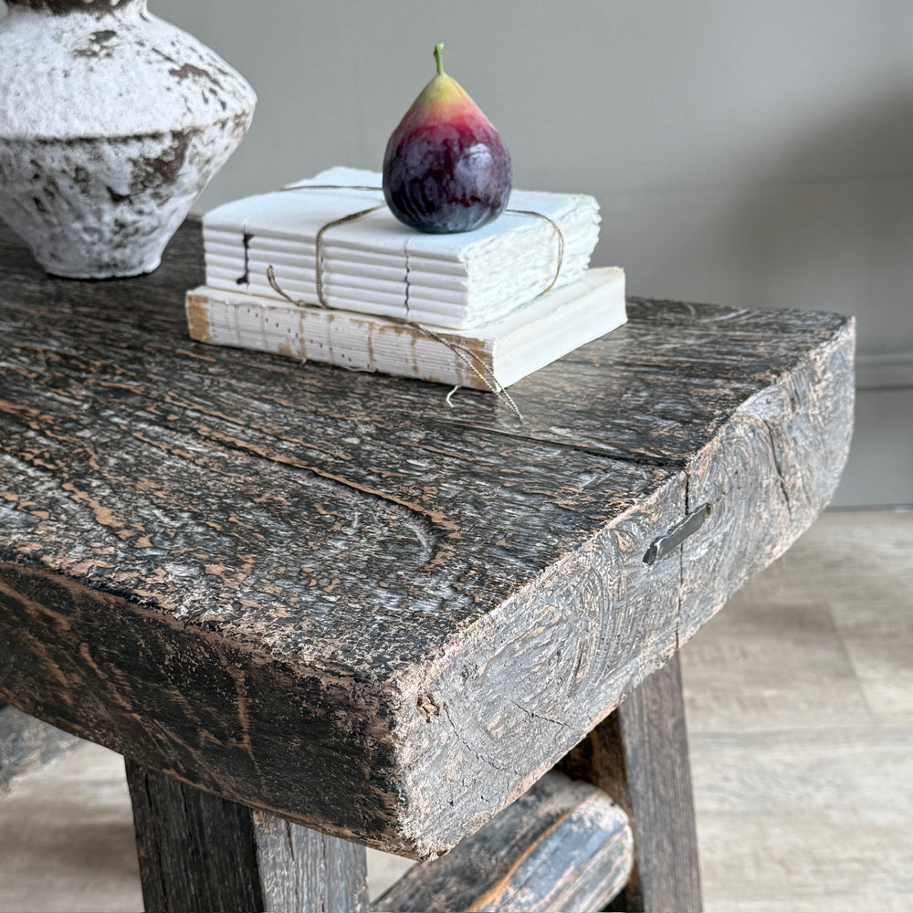 A rustic dark wood table or stool in a real interior setting, styled with a stack of white books tied with string and a purple fig on top, alongside a distressed ceramic vase.
