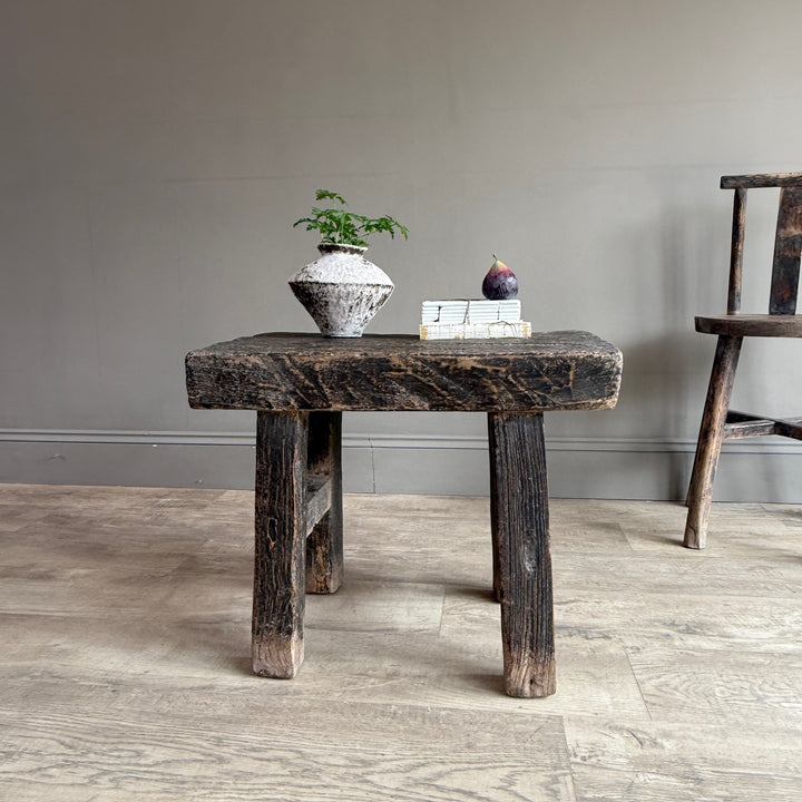 A rustic dark wood stool with a thick rectangular seat and four short legs, styled with a small potted plant and books on top, set against a neutral gray wall and light wood floor.