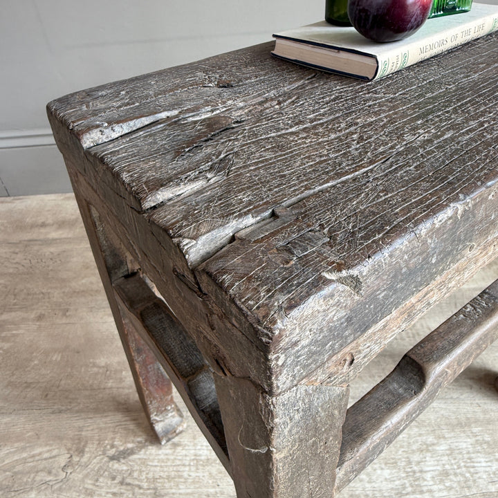 A rustic, weathered dark brown wooden stool with a thick, heavily textured tabletop and integrated side supports, shown in a home setting with a book and fruit on top.