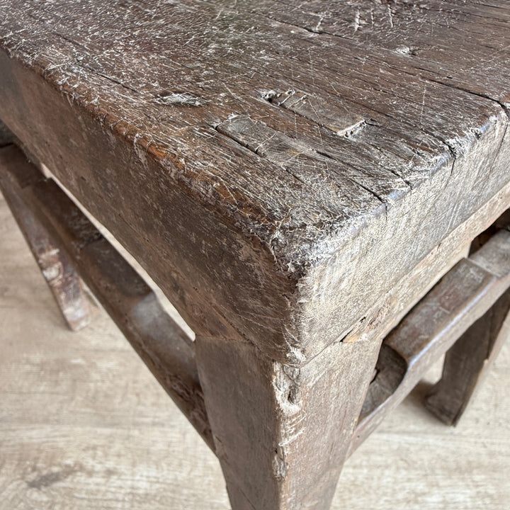 A close-up of a rustic, weathered dark brown wooden stool showing the textured seat and corner joint.