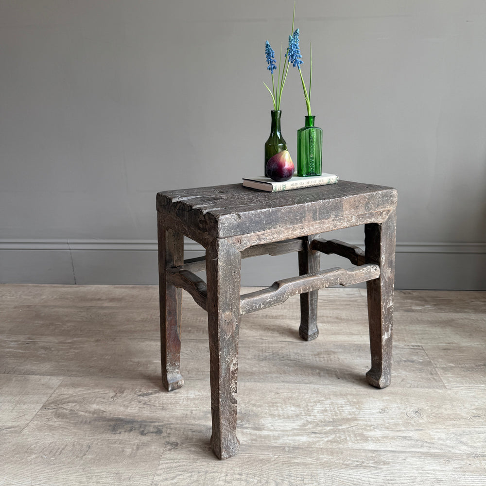 A rustic, weathered dark wood stool with a distressed finish, styled with two green glass bottles and a small book on top, set against a neutral gray wall and light wood floor.
