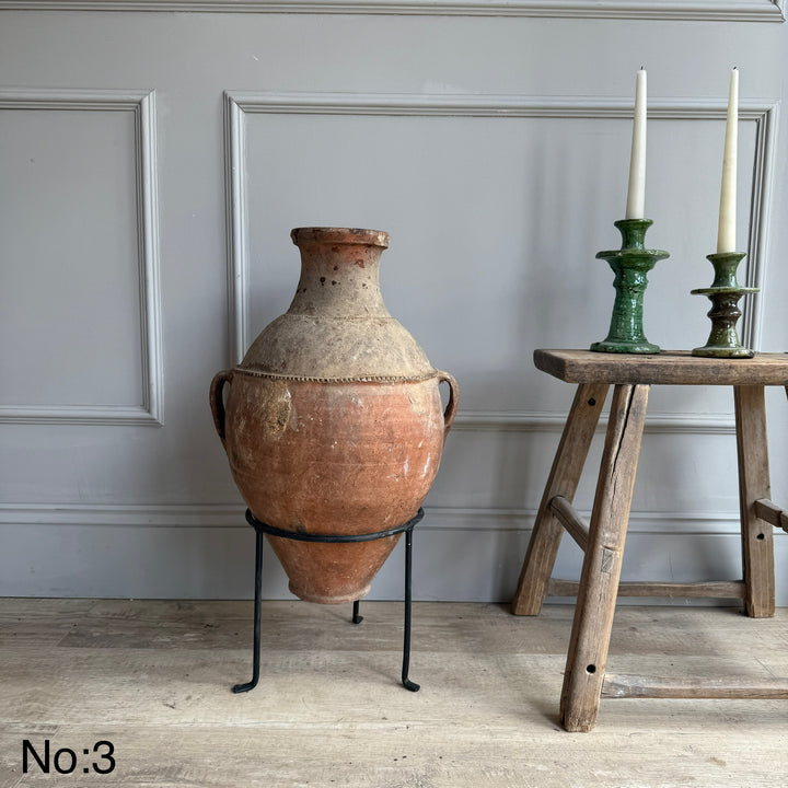 Large terracotta vase on a stand next to a wooden table with green candlesticks against a gray wall.