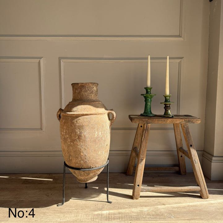 Large ceramic vase on a stand next to a wooden stool with candles against a neutral wall.