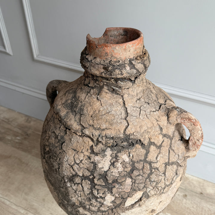Vintage ceramic jar with cracked surface on a wooden floor against a gray wall.