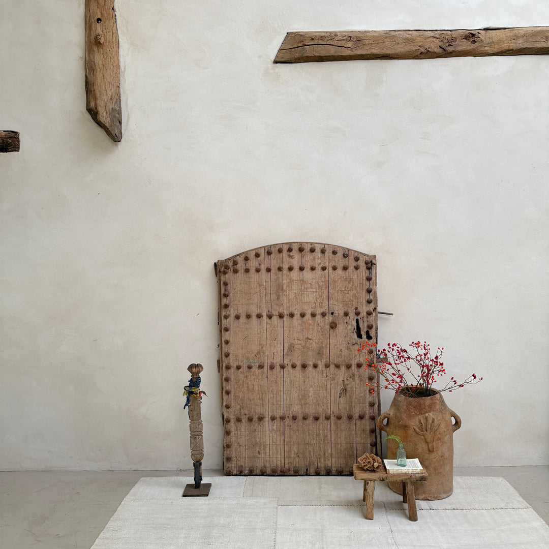 Large wooden door with decorative nails in a minimalistic room with a vase and plant.