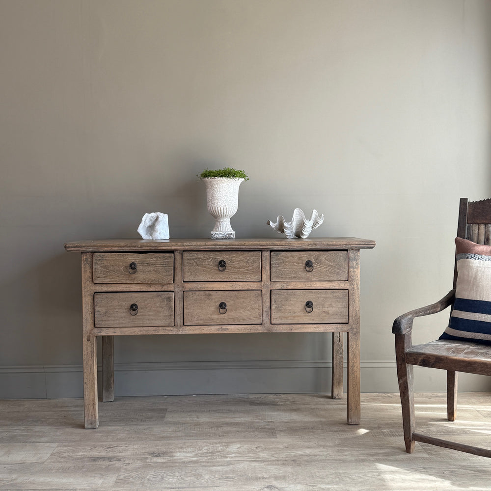 Wooden console table with decorative items against a plain wall, next to a chair.