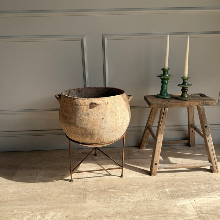Vintage pot on a stand next to a wooden stool with candle holders against a paneled wall.