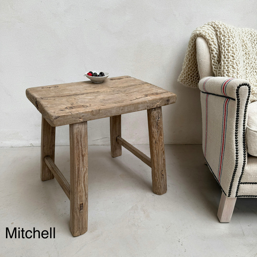 Wooden side table with a bowl of fruit next to a chair with a textured cover on a light-colored floor.