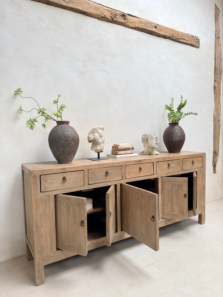 A light natural wood sideboard cabinet with multiple drawers and two cabinet doors, styled with two dark brown vases, small sculptures, and books against a textured off-white wall.