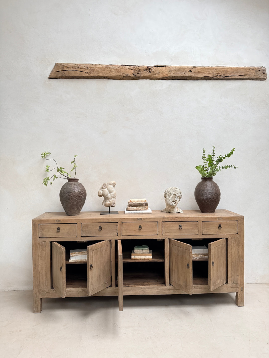 A light natural wood sideboard cabinet with multiple drawers and open cabinet doors, styled with two dark brown ceramic vases, small sculptures, and books against a textured off-white wall with a rustic wooden beam above.