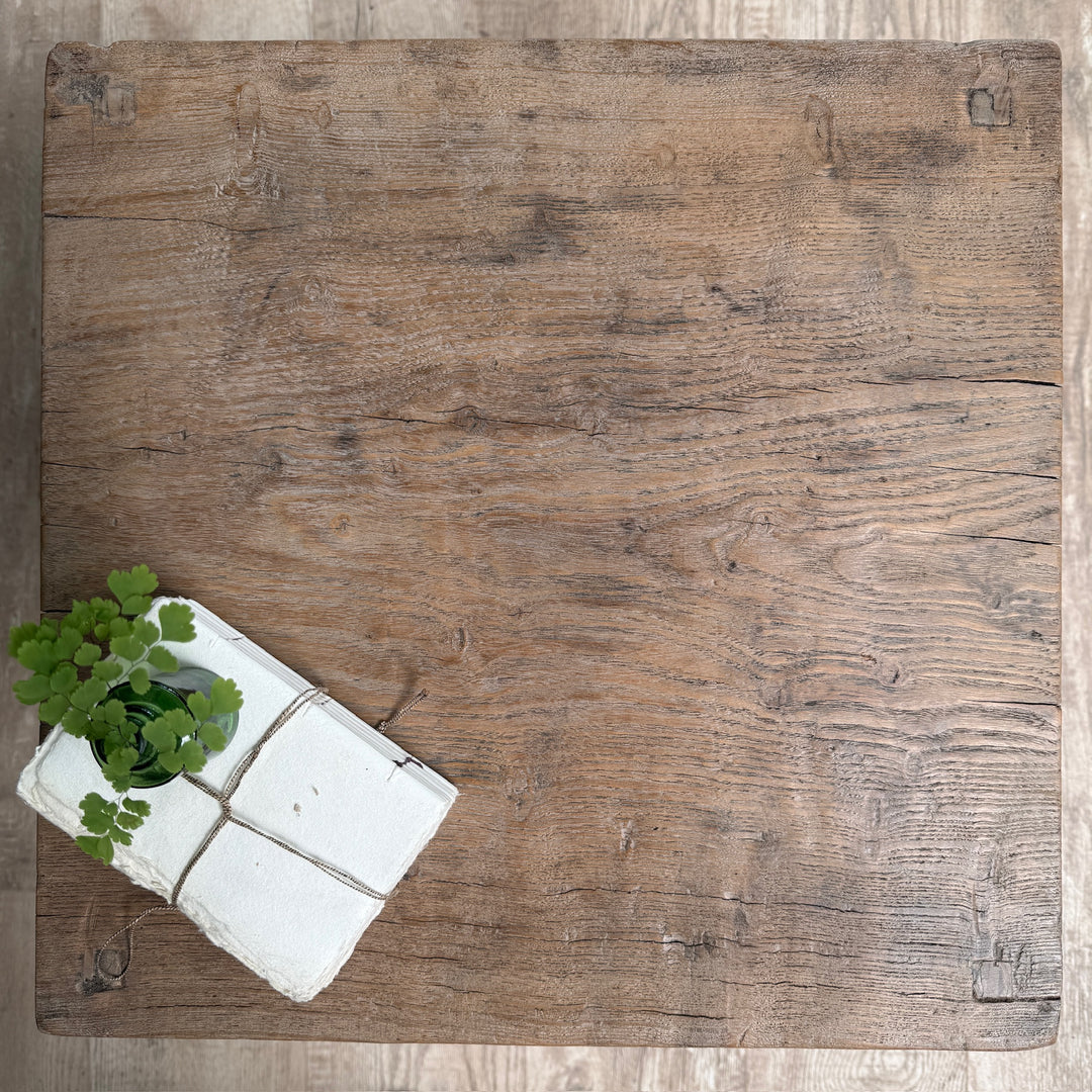 A square rustic wooden board or tray with a weathered brown wood grain finish, photographed from above on a light wood floor. A small white box with green leafy stems sits in the lower-left corner.
