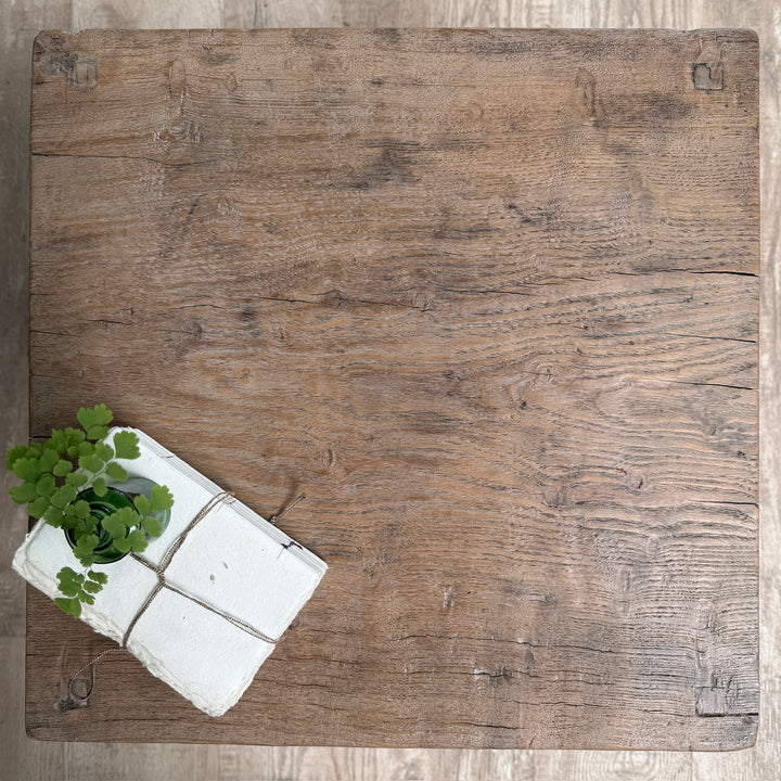 A square rustic wooden board or tray with a weathered brown wood grain finish, photographed from above on a light wood floor. A small white box with green leafy stems sits in the lower-left corner.