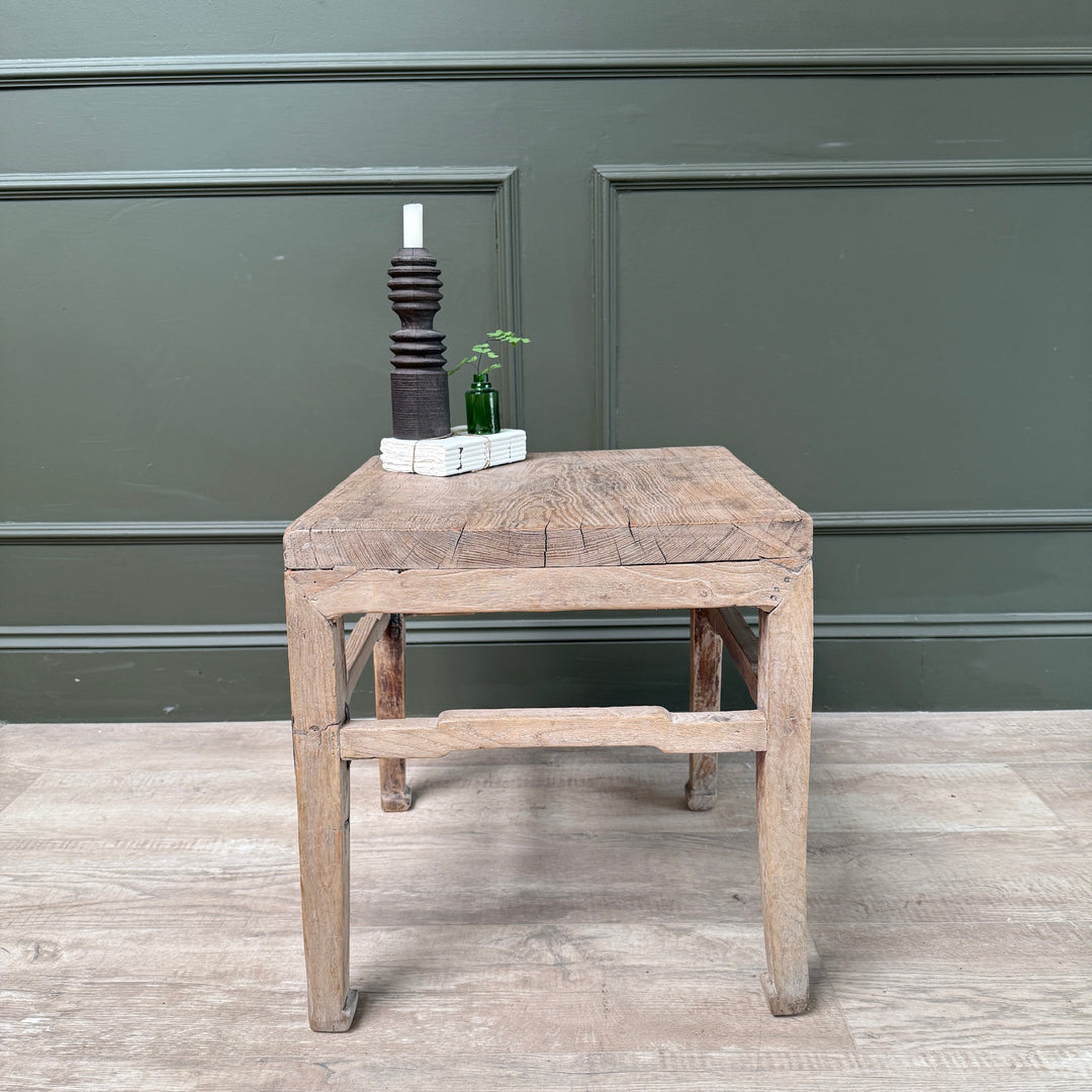 A rustic, square-shaped wooden side table with a weathered brown top and legs, styled with a dark wooden candle holder and a small green plant against a dark green paneled wall.