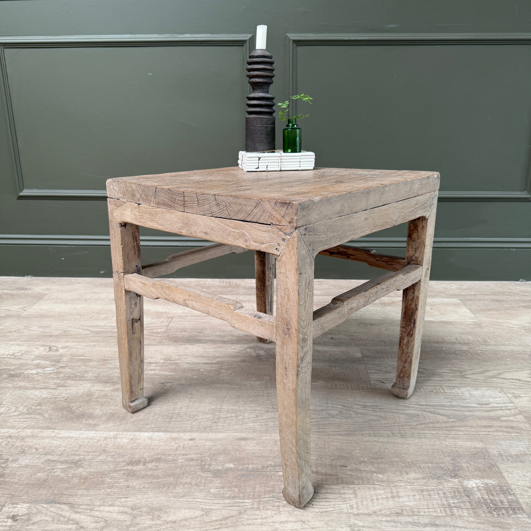 A rustic, square wooden side table with a weathered, light brown finish and distressed details, styled with a dark wooden candle holder and a small green plant on top, set against a dark green paneled wall and light wood floor.