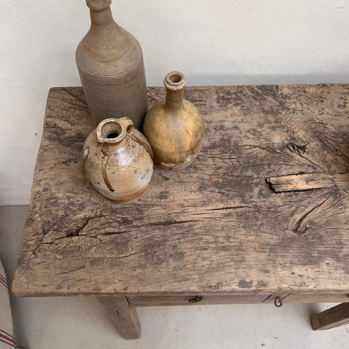 Three rustic handmade ceramic bottles and vases in earthy beige, brown, and gray tones arranged on a weathered wooden table against a plain off-white wall.